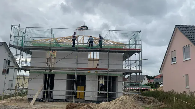 Rohbau eines Massivhauses mit Gerüst, Richtkranz und Arbeitern auf dem Dachstuhl in einem Wohngebiet.